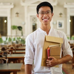 young-smiling-asian-male-student-eyeglasses-with-notepads-joyfully-looking-camera-library-university